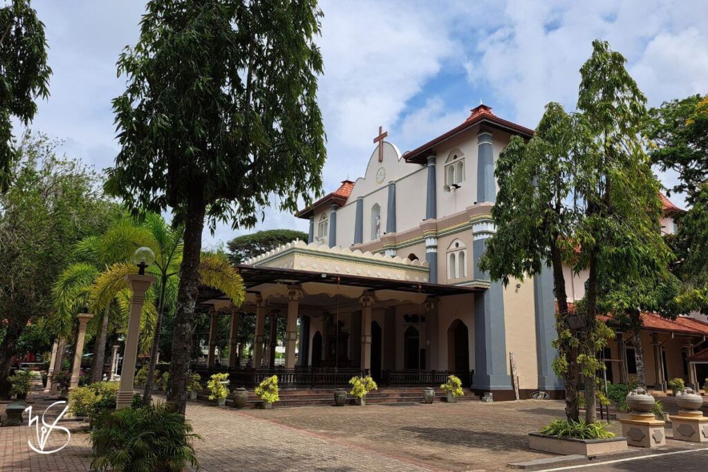 A photo of the Katuwapitiya Church. The building is painted in cream and a bluish grey, with a large cross adorning the roof of the church. Numerous trees are scattered around the church, providing shade from the sunlight.