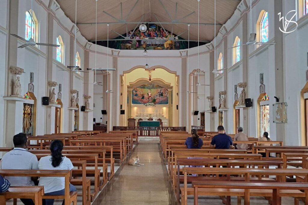 The interior of the renovated church. The faithful sit on wooden pews, some of whom are kneeling. A dog rests sleepily in the aisle. Stone statues and stained glass windows adorn every corner of the church, and a mural depicting the Last Supper is painted at the top of the roof. A statue of St Sebastian is atop the altar with a backdrop of painted angels in Heaven.