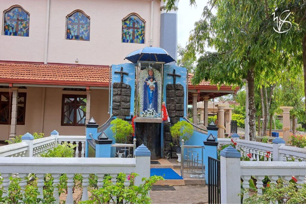 A photo of a shrine on the church grounds, dedicated to the Virgin Mary. Enclosed in a stone fence and surrounded with plants, the shrine is painted blue with a small covering to protect the statue from the rain. The Virgin Mary stands, encased in glass, bearing the infant Jesus in her arms.