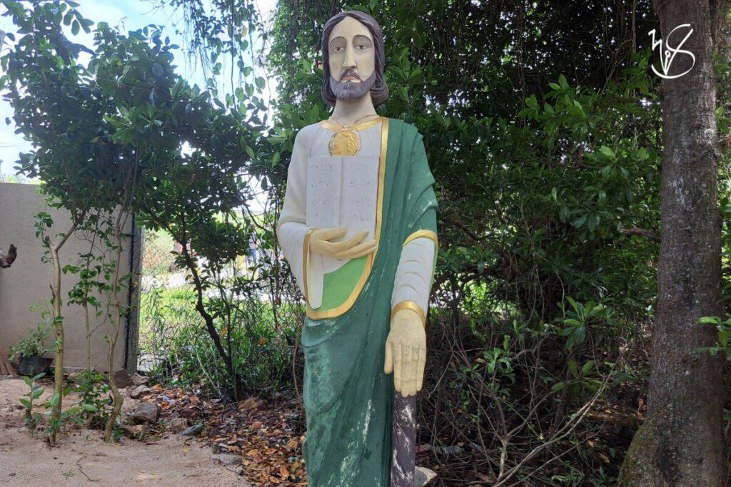 Photograph of the statue of St. Jude at the entrance of the Kurulukele Church. The statue is garbed in green and white robes that are accented with gold. The statue itself is rather worn at the bottom, perhaps from the flooding of the nearby lagoon. St. Jude bears a stern look on his face, and a club in one of his hands with a book in the other. The statue is surrounded by wild trees and greenery.