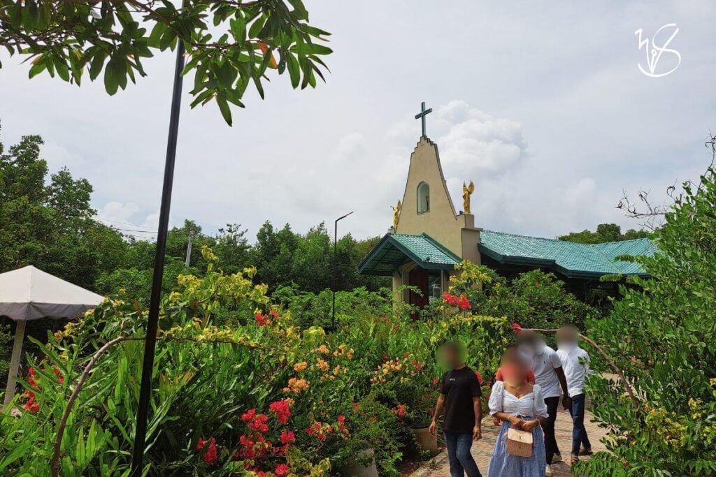 A photograph of the exterior of the Kurulukele Church. The church is painted in a yellowish cream with a green roof, and statues of golden angels perch on either side of the cross that is placed at the highest point of the roof. In this particular photo, the pathways in which flowers and plants grow is the focus, and the church blends seamlessly with the greenery against the cloudy skies.