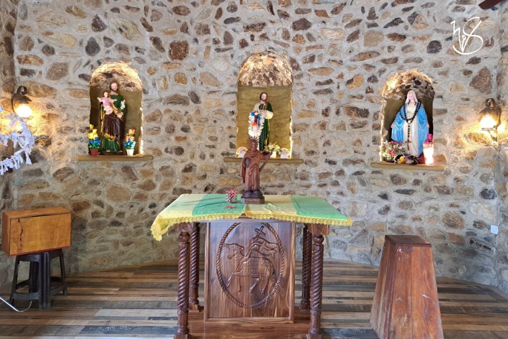 A photograph of the interior of the chapel, wherein statues of Jesus Christ, Mother Mary and St. Jude beckon to the faithful. Apart from the stone walls, the floor, mite box, and other furniture within are made of wood.