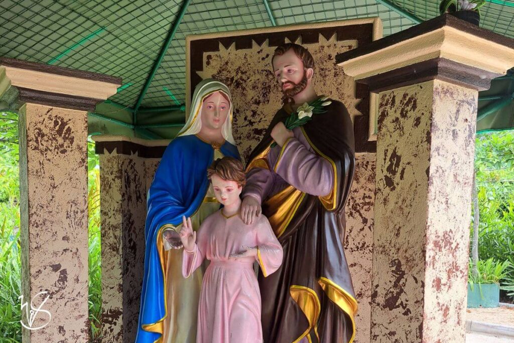 Photograph of one of the gazebo-like complexes in the Kurulukele complex. A statue of the Holy Family lies here beside pillars. St Joseph, garbed in brown and gold, has his hand on the young Jesus' shoulder, bearing a thoughtful look on his face, whereas the Virgin Mary, garbed in blue and white, has her palm outstretched, directing devotees' attention to the young Jesus, who dons pink robes.