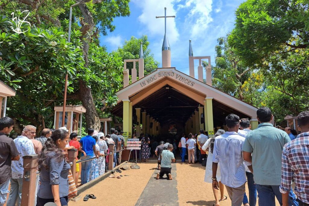 A photo taken at the entrance of the Marawila Church. Crowds of people are queuing on opposite sides of this small church, and devotees are kneeling on a path of cement for penance. The church is painted in a faint, almost pastel orange, and the words In Hoc Signo Vinces are presented on the exterior in silver lettering (which translates as In this sign thou shalt conquer). A steel cross adorns the top of the church spire. The skies are a clear shade of blue with minimal clouds, and the trees grown around the church provide shade to the faithful.