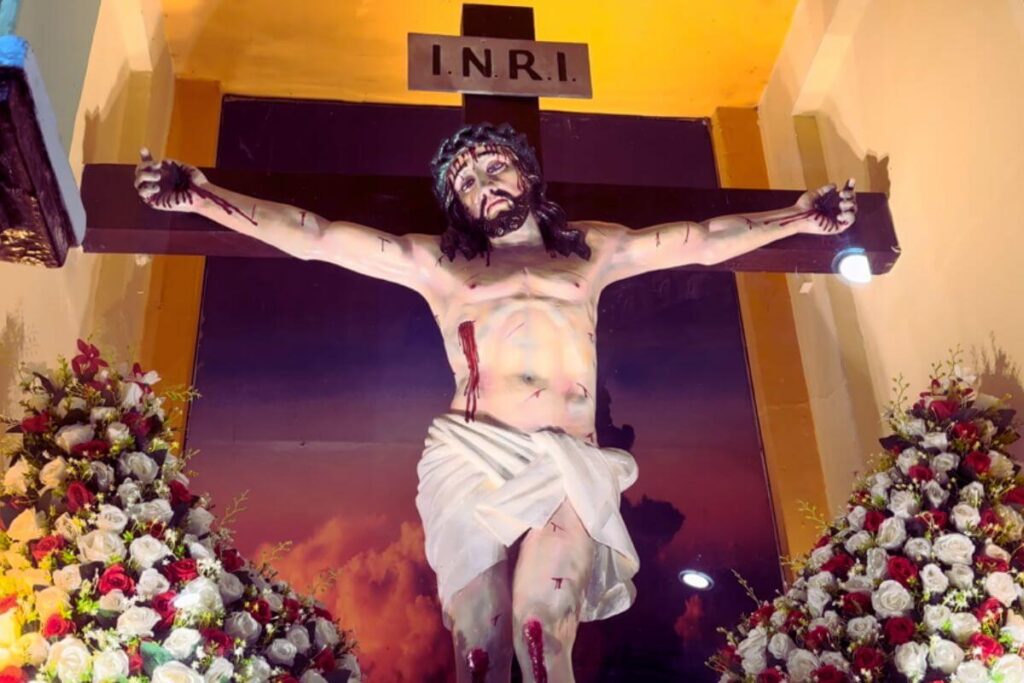 Statue of the crucified Christ, described as above. Blood gushes from the wounds on His hands, knees, and side, and a photo of the clouds forms the backdrop for the wooden cross. On either side of the statue are plastic roses of white and red.